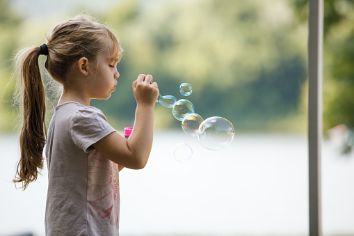 Profile view of adorable little girl standing outside and having fun blowing soap bubbles.