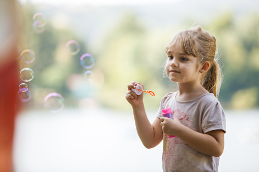 Copy space shot of adorable little girl standing outside and having fun by herself. She is blowing soap bubbles and watching them fly away.