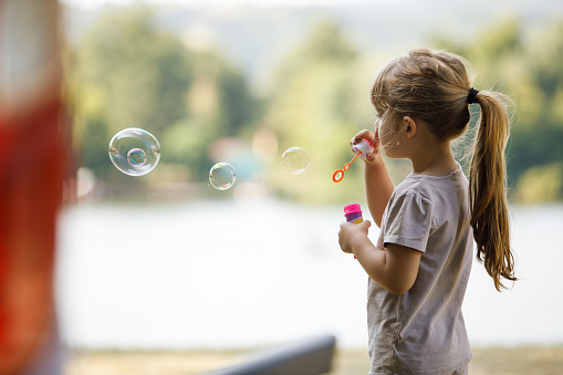 Copy space shot of adorable little girl standing outdoors and playing with soap and blowing bubbles with a wand.