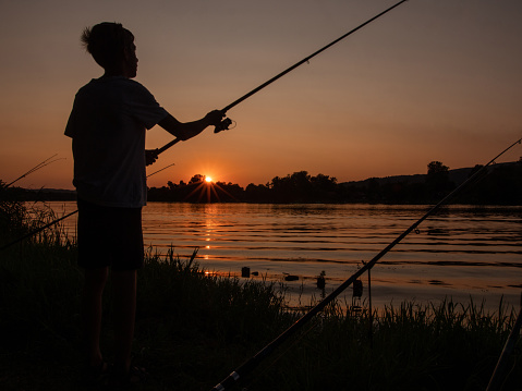Wide shot of teenage boy standing on the riverbank and throwing a bait in the water while fishing during a beautiful sunrise or sunset.
