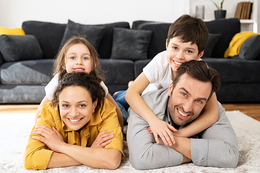 Happy multiracial family of four laying at the floor in cozy living room, parents and kids laughing while embracing with each other. Family feeling happy during the weekend