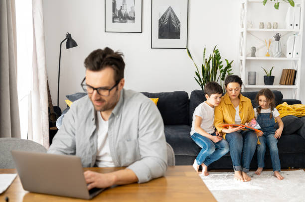 istock Man using laptop for freelance work sitting at the table at home while family sitting peacefully together on the sofa. Happy father wearing glasses is working 1391474190