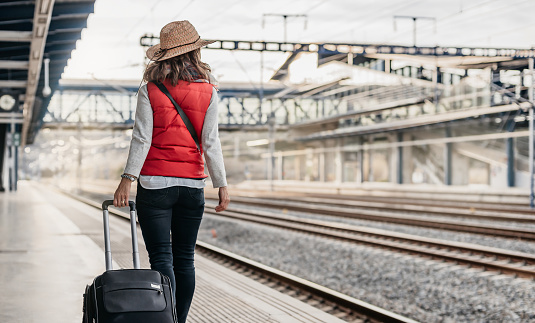 medium shot of a tourist in a hat and red vest carrying a suitcase and walking through the station waiting for the train. her back is turned. concept of travel and vacation.