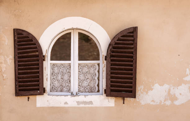 Two old wooden windows sit on a faded wall, revealing lace curtains. One window has a noticeable hole, showcasing the building's age and neglect over time