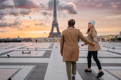 Happy couple, woman with long blonde hair wearing a white hat and beige jacket with brown dots, man with short brown hair and beard, wearing a beige coat, running on Parvis des Droits de l'Homme, Paris, in the direction of Eiffel Tower, woman smiling at her boyfriend, twilight with sunset and Eiffel Tower in the background, focus on forefront, horizontal