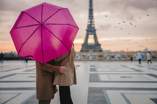 Couple, man and woman kissing each other at Parvis des Droits de l'Homme, Paris while hiding behind a pink umbrella, Eiffel Tower in the background, focus on forefront, evening with sunset sky, horizontal