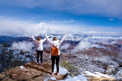 Joyful young women enjoying life outdoors. Friends standing on top of the mountain with arms raised. Happy friends hiking on winter vacation in the mountains.  Grand Canyon National Park, Arizona, USA.