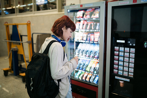 Side view of a young happy Caucasian woman waiting for her train on a subway station, looking at a vending machine and choosing a refreshment while she waits