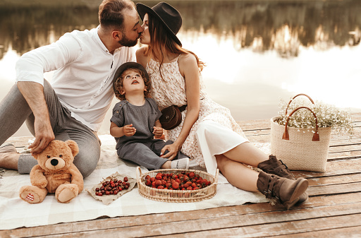 Loving couple kissing and sitting on blanket with little child while having picnic with fresh berries on wooden quay near pond in summer