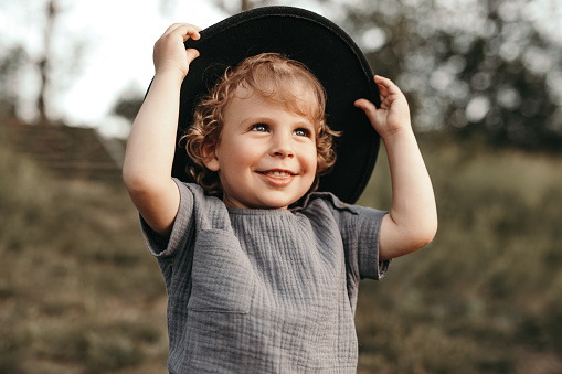 Adorable curly haired toddler boy in summer wear and hat smiling happily while having fun in nature