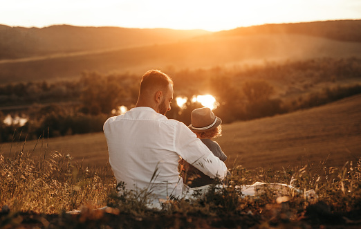 Back view of loving dad embracing little son while sitting together on hill slope and enjoying time, together during summer adventure in countryside at sunset time