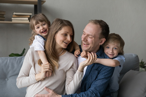 Happy young man touching belly of smiling pregnant wife, spending time with joyful adorable children siblings, resting together on cozy sofa. Loving family waiting for third baby, relations concept.