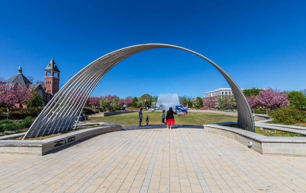 Rock Hill, SC, USA-10 April 2022: The Fountain Park in downtown. Spring day, with sunny blue sky.  Focus on artistic steel arch, with two adults, two children.  Fountain in background.
