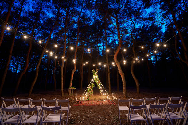 istock Festive string lights illumination on boho tipi arch decor on outdoor wedding ceremony venue in pine forest at night. Vintage string lights bulb garlands shining above chairs at summer rural wedding 1391544376