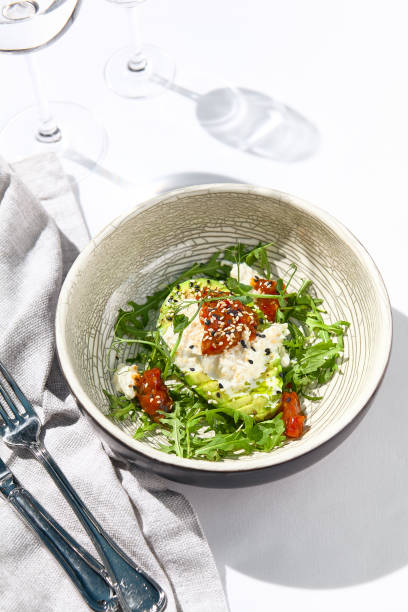 istock Avocado and stracciatella cheese salad. Fancy dinning with Italian salad bowl on white table with simple contemporary decor. Sunlight and harsh shadow still life. Avocado, cheese and tomato salad. 1391549701