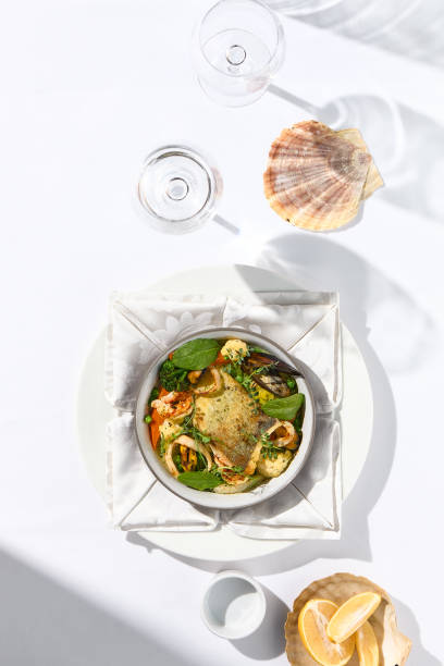 istock Elegant seafood stew with cod fish and vegetables in pot. Healthy fish dish - baked codfish, shrimp, mussels on broccoli and tomato. Serving white tablecloth and glass in summer day with shadow. 1391549744