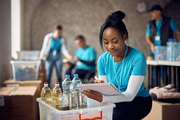 Black woman working as volunteer and making list of donated groceries for food bank. African American female volunteer making list of food supplies while working at humanitarian aid center. altruism stock pictures, royalty-free photos & images