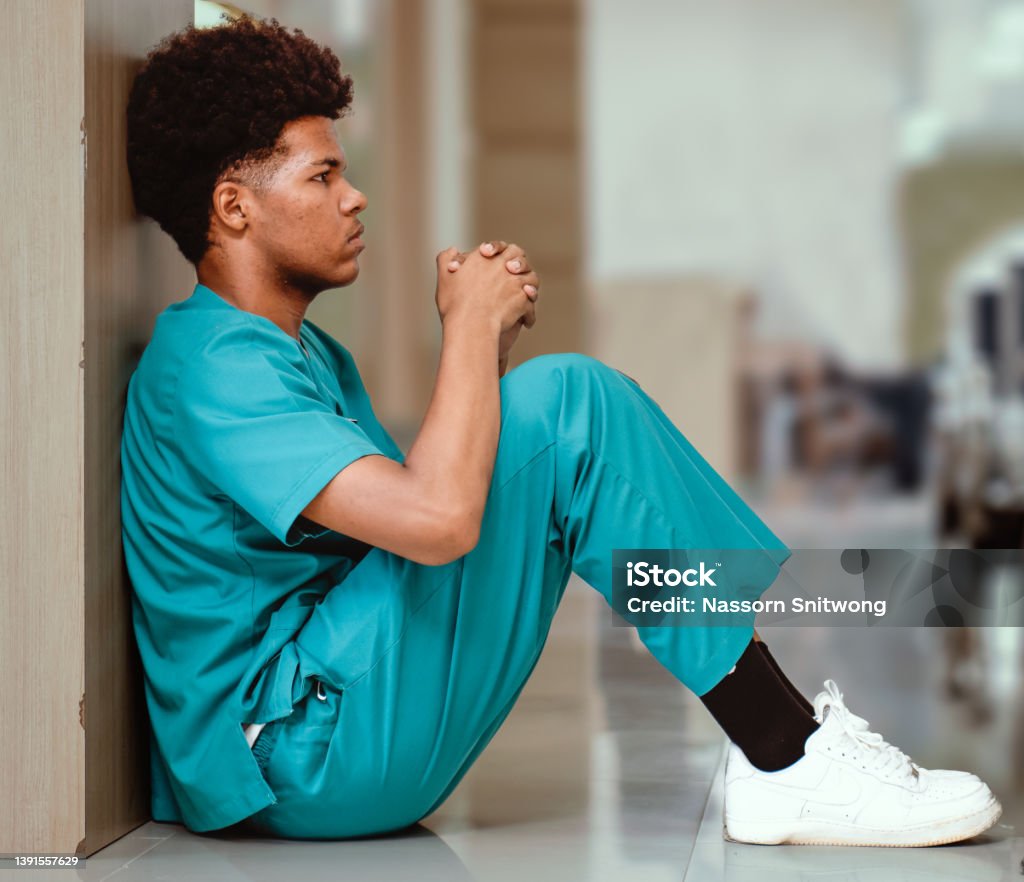 Emotional stressed intern doctor sitting against the wall on floor in a hospital corridor. Emotional stressed intern doctor sitting against wall on floor in hospital corridor. Distraught medical student in seriously depressed, sad or exhausted. Overworked male nurse sits in despair or tired Medical Student Stock Photo