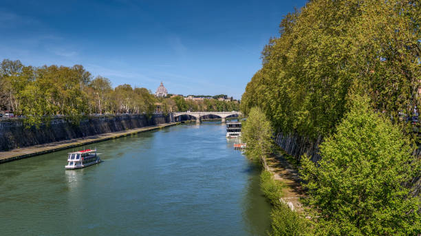 A tourist ferry sails along the Tiber River near the Trastevere district in the heart of Rome A tourist ferry sails along the Tiber River near the Trastevere district, in the historic heart of Rome. Along the horizon stands the majestic dome of St. Peter's Basilica and the Giuseppe Mazzini Bridge. On the sides the high walls of the embankments built at the end of the 18th century, surmounted by secular trees. In 1980 the historic center of Rome was declared a World Heritage Site by Unesco. Wide angle image in 16:9 and high definition format. international match stock pictures, royalty-free photos & images