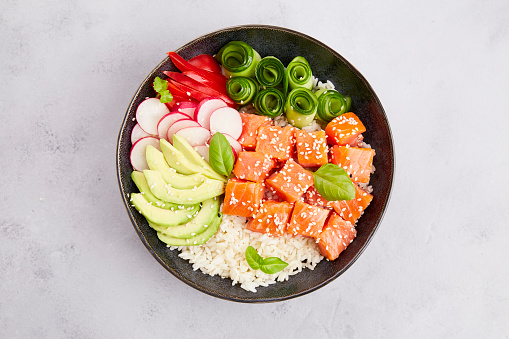 Poke bowl with salmon tuna, rice, avocado, cucumber, radish, pepper, sesame seeds on grey background. Close-up. Hawaiian diet food with fish, pokebowl. Top view.