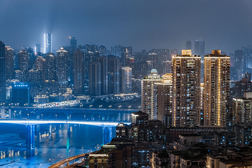 2021, June 5th, Chongqing China - The city at night, in the foreground we see a building illuminated in yellow and to the left we see an illuminated bridge and the yellow river flowing underneath it. In the background we see what appears to be an endless amount of illuminated buildings spreading.