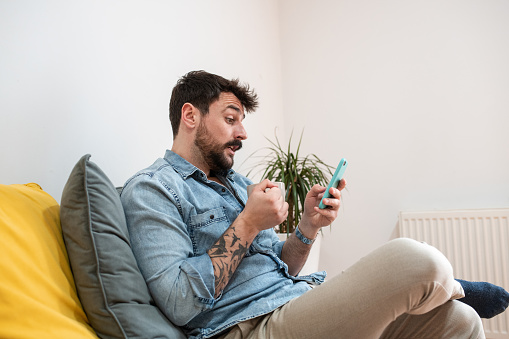 Handsome man drinking morning cup of coffee and reading the news. Young tattooed man with a beard sitting in his living room.
