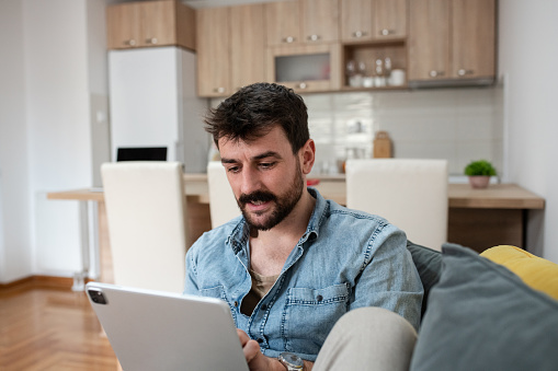 Handsome man with a beard and tattoos on his arm, holding a tablet device - iPad. He is sitting on the sofa and reading the news.