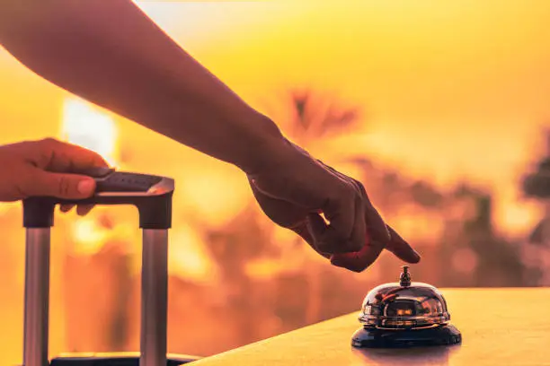 Photo of Woman with suitcase ringing hotel service bell with sea and palm tree view on sunset. Travel concept. 24-hour beach hotel front desk. Late check-out.