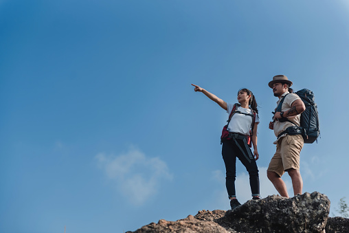 An Asian backpacker trekking man and woman are reaching the top and pointing to the goal as a concept of tourism and teamwork.