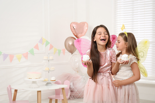Cute little girls wearing fairy costumes with delicious cupcakes in decorated room. Space for text