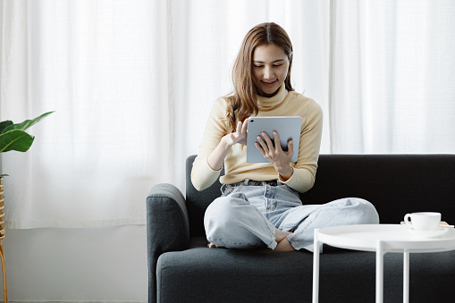 A woman plays a tablet on the sofa in her living room at home, she is resting on weekends after a hard day's work, she relaxes by watching movies and listening to music on her tablet. Holiday concept.