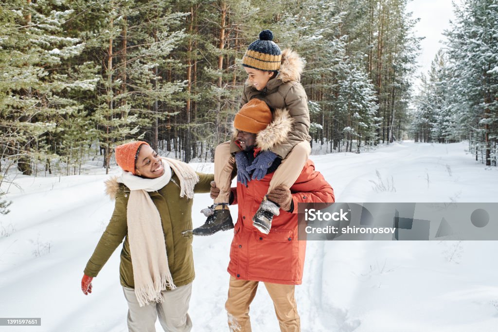 Young contemporary family of father, mother and son enjoying winter day in park Young contemporary family of father, mother and son enjoying winter day in park while African American man carrying little boy on shoulders Snow Stock Photo