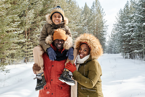 Cheerful family of three in warm winterwear spending day in winter forest or park among firtrees and pines covered with snow