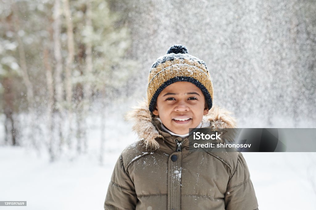Cheerful little boy of African ethnicity wearing warm winter jacket and beanie Cheerful little boy of African ethnicity wearing warm winter jacket and beanie hat while having fun in the forest on frosty day Child Stock Photo
