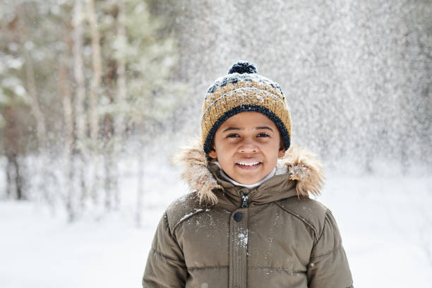 Cheerful little boy of African ethnicity wearing warm winter jacket and beanie Cheerful little boy of African ethnicity wearing warm winter jacket and beanie hat while having fun in the forest on frosty day children in winter stock pictures, royalty-free photos & images