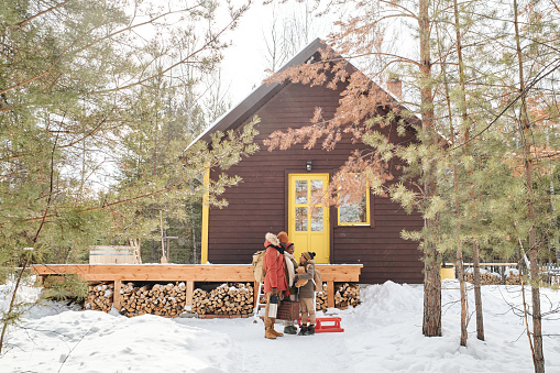 Contemporary family of parents and little son in winterwear standing by their country house located in deep woods among pinetrees