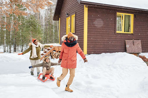 Young cheerful black man having fun with his family while pulling sledge with happy cute boy outstretching his arms and enjoying ride