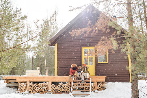 Happy family of three in winterwear having chat on wooden porch by their country house or cottage while spending vacation there