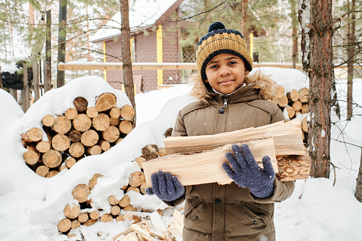 Cute little boy in warm winter jacket and knitted beanie holding firewood from woodpile while standing against country house of his family