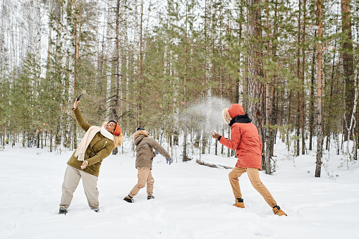 Young African American female in winterwear making selfie with her son and husband playing snowballs in the forest on winter day