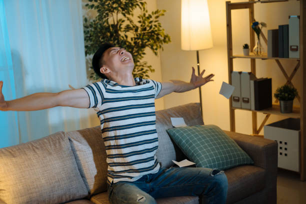 istock happy successful young Asian billionaire man smiling happily open arms stretching and looking up. Wealth investment economic concept. cheerful male sitting in home living room couch win lottery 1391601251