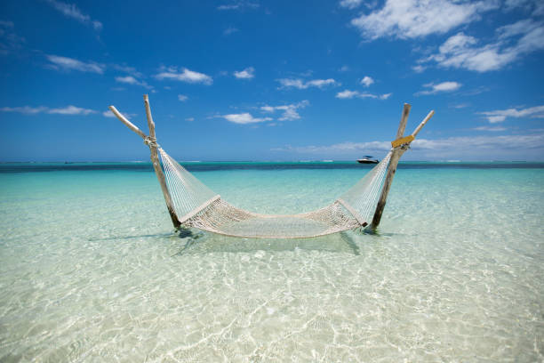 A woman relaxes in a colorful hammock suspended above shallow turquoise water on a sunny tropical beach. The scene captures a peaceful vacation moment with blue skies, calm waves, and a carefree atmosphere, perfect for themes of summer, travel, leisure, and coastal escapes.