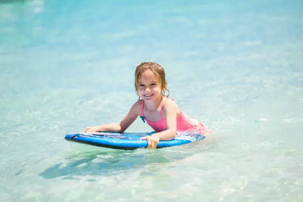 Photo of Little baby girl - young surfer with bodyboard has a fun on small ocean waves. Active family lifestyle.