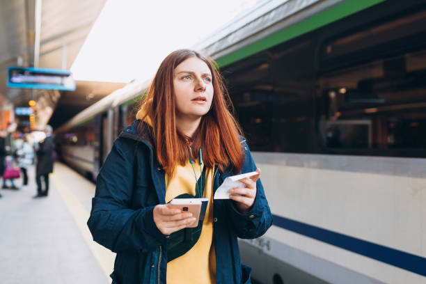 istock Young redhhead woman waiting on station platform with backpack on background electric train using smart phone. Railroad transport concept, Traveler 1391608877