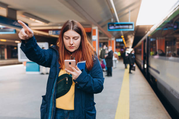 istock Young redhhead woman waiting on station platform with backpack and pointing finger on background train using smart phone. Railroad transport concept, Traveler 1391610198