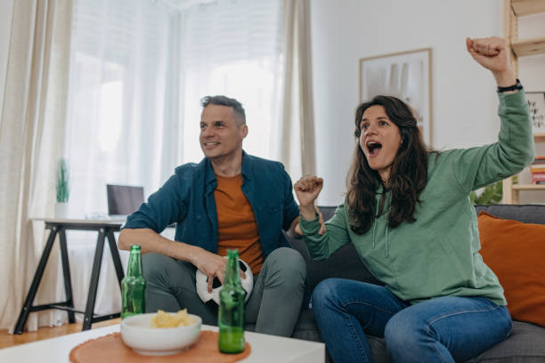 Hope they will win Husband and wife passionately cheering for their football team at home, men holding ball and woman is  loud trophy wife stock pictures, royalty-free photos & images