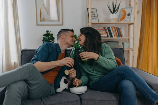 A kiss of victory Happy couple sitting on sofa at home watching game, holding bottle of beer in one hand and celebrate victory of their football team trophy wife stock pictures, royalty-free photos & images