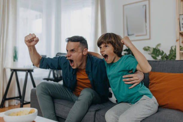 Boy watching soccer match with father at home, sitting on sofa, cheering very loud