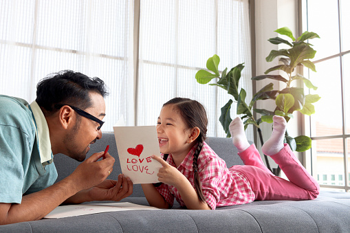 Happy Father's Day, adorable cute little girl with young man dad lying on sofa in living room, making handmade greeting card for calibration. Happy family daughter and father spending time together.