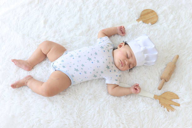 istock Six months crawling baby kid sleeping next to kitchenware and wooden rolling pin on fluffy white rug, wearing chef hat, costumes like a chef, childhood and dreaming career concept. 1391641840